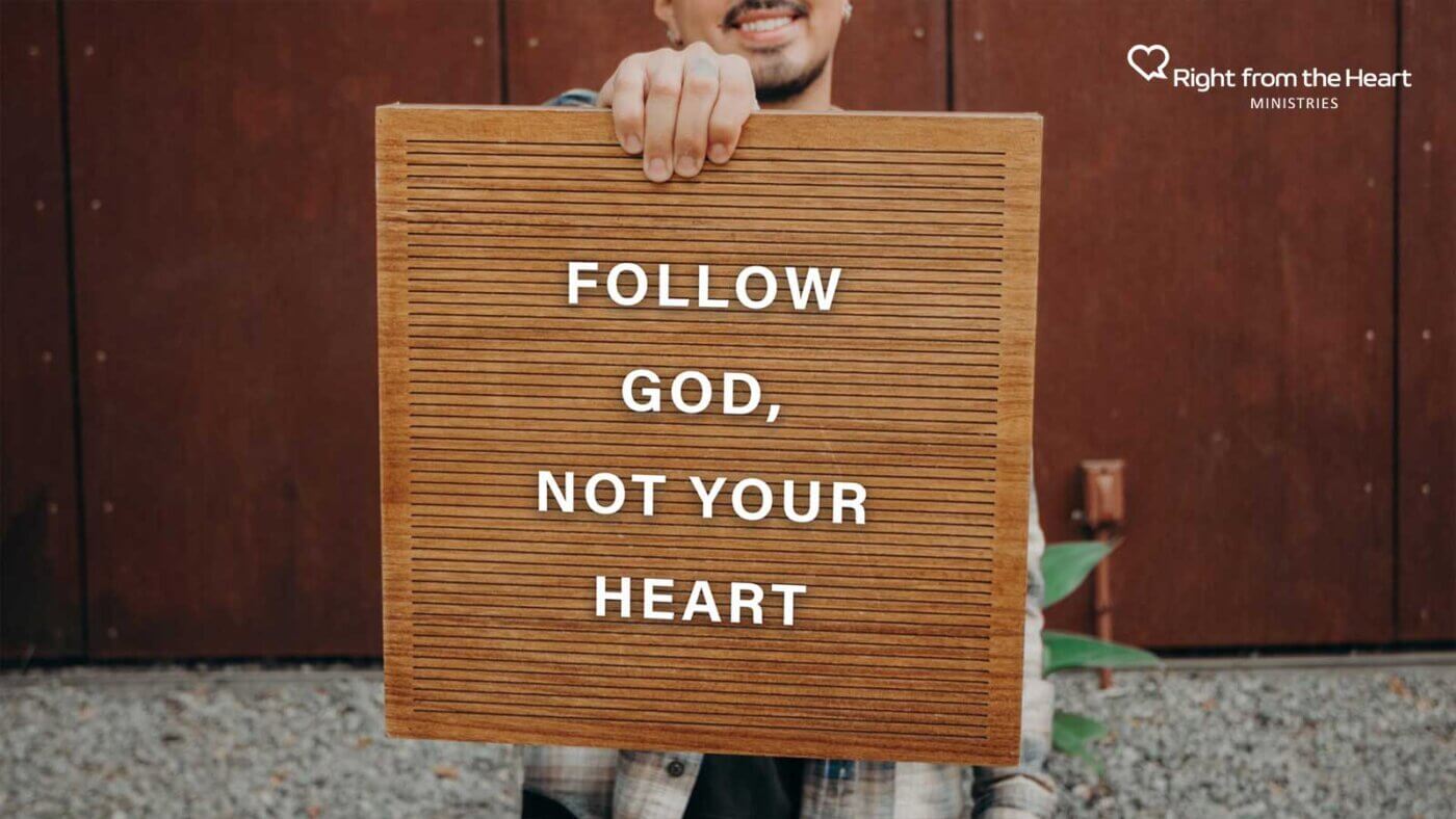 Person holding a wooden sign that reads 'FOLLOW GOD, NOT YOUR HEART' with a ministry logo in the top-right corner.